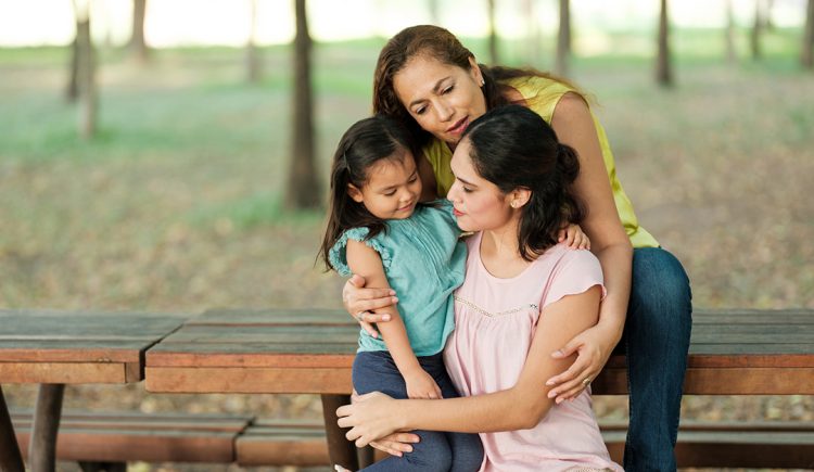 three generations of latina women sit on a bench