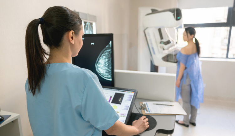 a woman gets a mammogram as a tech looks at imaging