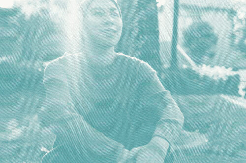 A younger Asian woman wears a headscarf and looks away from the camera, sitting under a tree